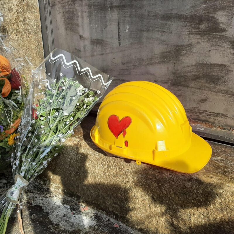 Mazzi di fori e un casco di protezione giallo, con disegnato su un lato un cuore rosso che sanguina, su di uno scalino di una porta d'ingresso della stazione di Brandizzo